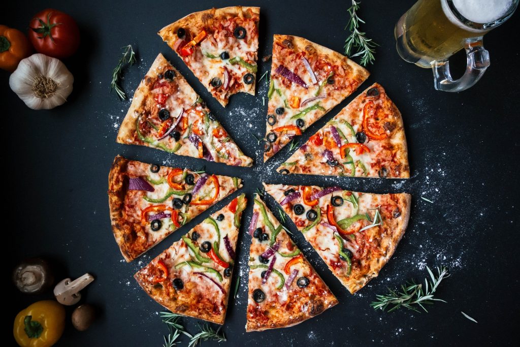 aesthetic ariel shot of a whole pizza topped with various vegetables, cut into slices that are slightly spread apart but still in a circle. Pizza is sitting on a black table with some flower and rosemary sprinkled around it. Edges of the screen have various vegetables such as mushrooms, bell peppers, garlic, and a beer.